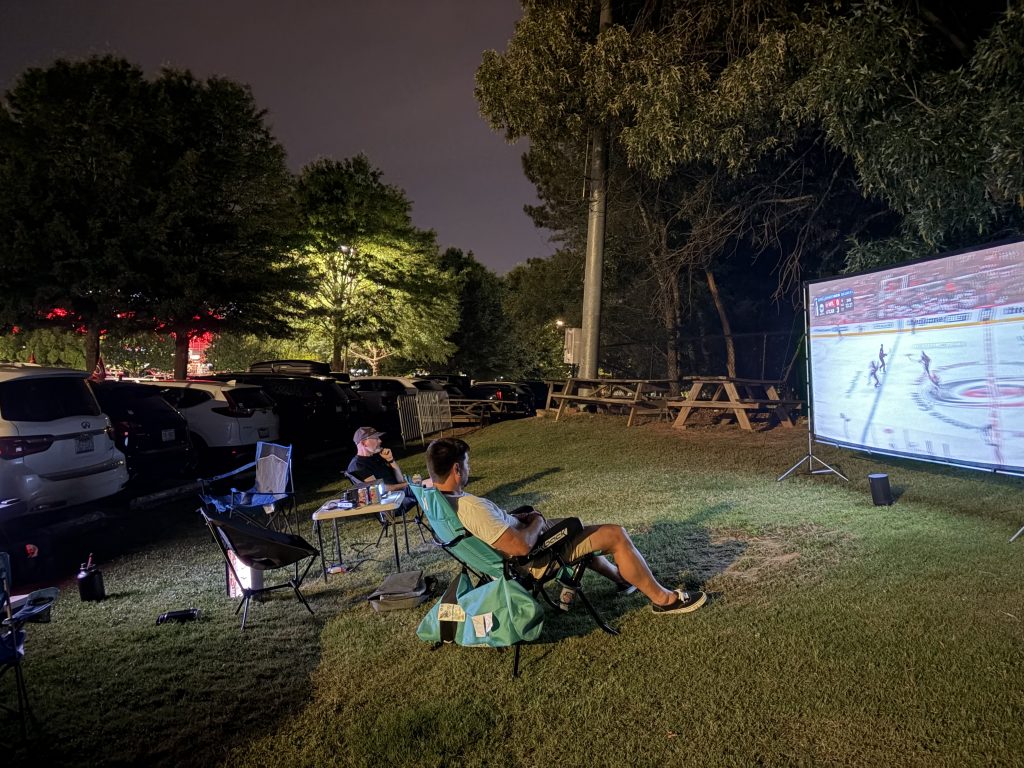 Two fans watching a Carolina Hurricane's game on a projector while sitting on lawn chairs in the grass.