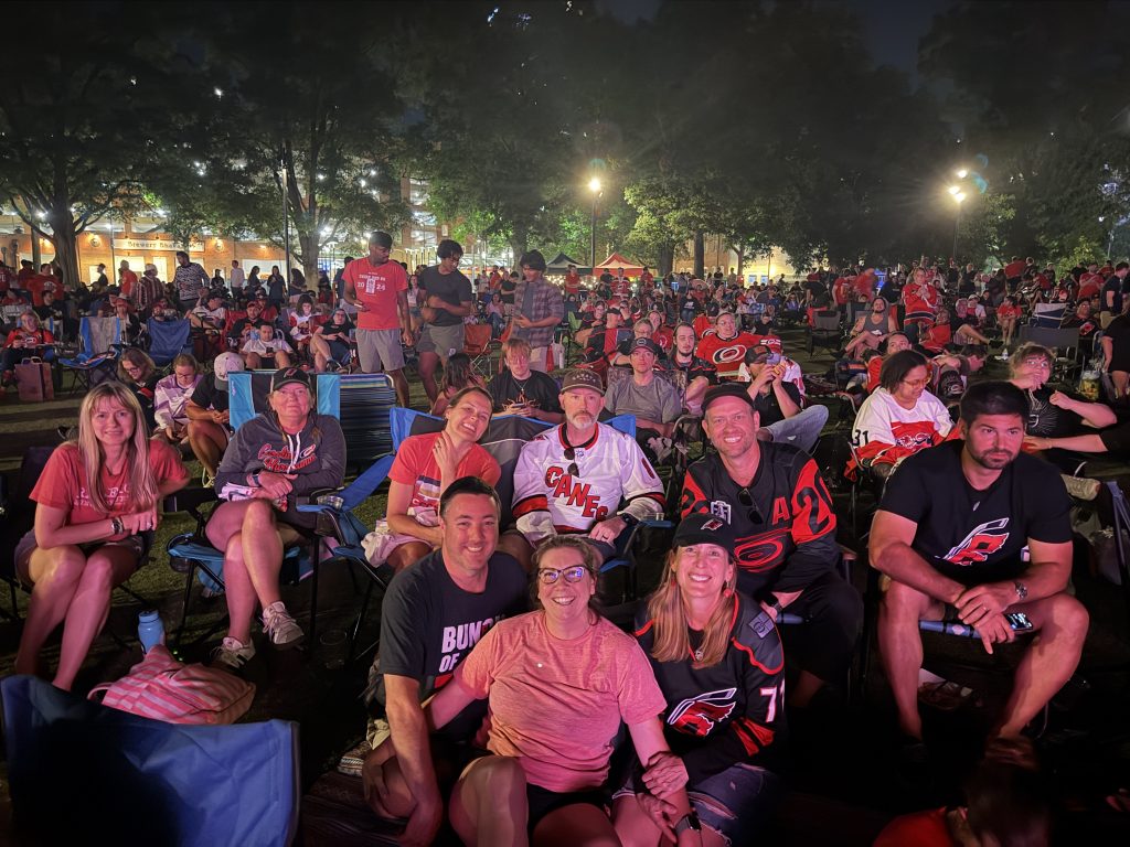 Hundreds of fans gather to watch the Carolina Hurricane's game while sitting in lawn chairs on a grassy field.