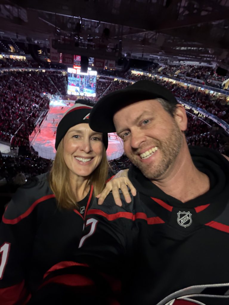 Husband and wife take a selfie in the stands during a Carolina Hurricane's game with a view of the rink behind them.